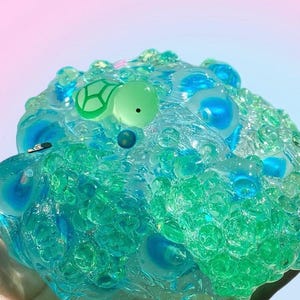 May include: A close-up of a clear, textured slime toy with blue and green hues. The slime has a turtle-shaped charm with a green outline and a black dot. The background has a gradient of pink and blue.