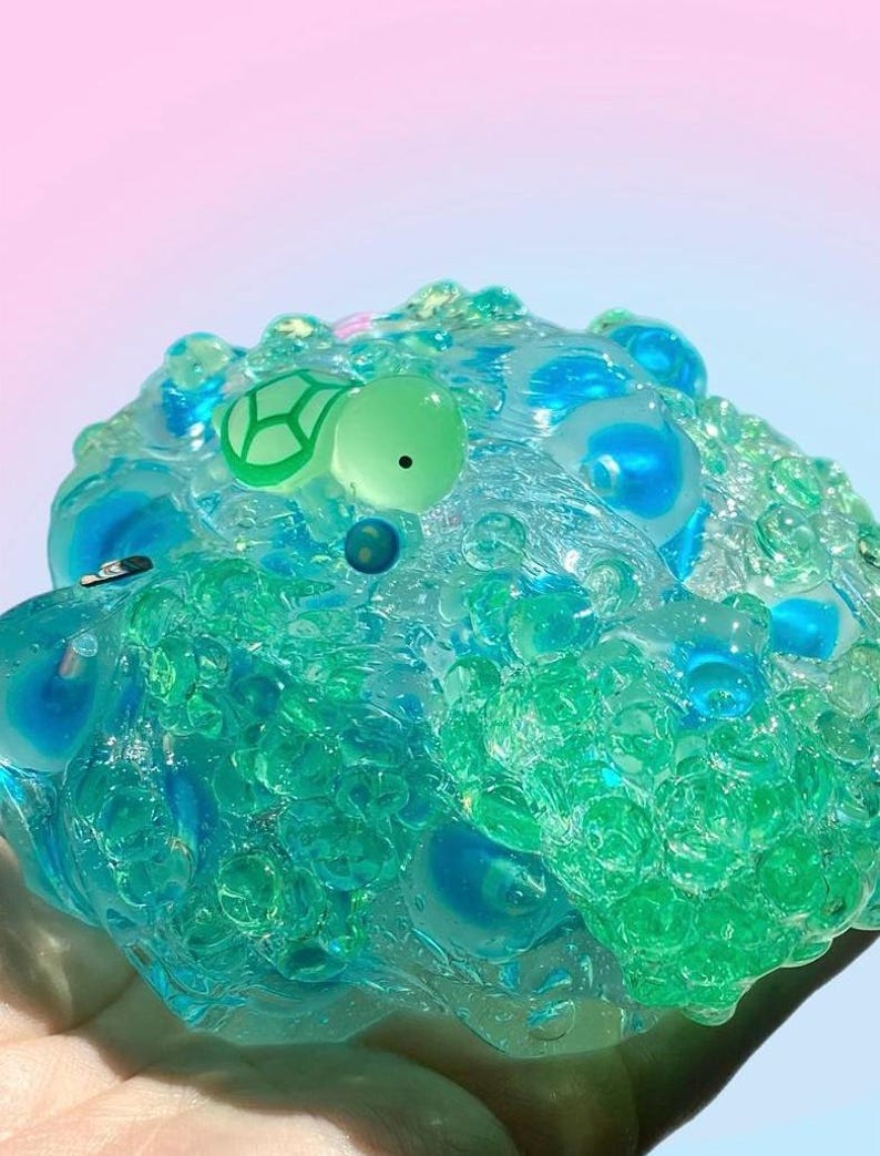 May include: A close-up of a clear, textured slime toy with blue and green hues. The slime has a turtle-shaped charm with a green outline and a black dot. The background has a gradient of pink and blue.