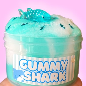 Peut inclure: Un récipient en plastique transparent rempli de slime bleu clair et blanc. Un petit requin en gomme bleu et blanc est placé sur le slime. Le récipient porte une étiquette indiquant "GUMMY SHARK".