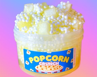 May include: A clear plastic container filled with yellow slime and white foam balls. The container has a blue label that says "Popcorn Crunch".