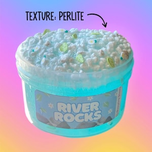 May include: A clear plastic container filled with light blue slime, topped with white perlite, and decorated with small green leaf and white flower-shaped embellishments. The container has a label that reads "River Rocks". The text "Texture: Perlite" is above the slime.