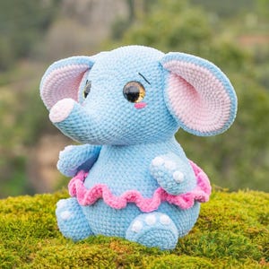 Puede incluir: Una muñeca de elefante de ganchillo azul hecha a mano con detalles en rosa en las orejas, la trompa y una falda con volantes. El elefante tiene ojos grandes y expresivos y almohadillas blancas. El peluche está sentado sobre un lecho de musgo verde.
