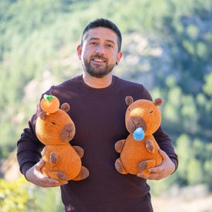 Puede incluir: Dos peluches de capibara naranja de ganchillo. Uno tiene una pequeña fruta naranja en la cabeza, el otro una bola azul en la boca. Los juguetes tienen detalles marrones y los sostiene una persona con un suéter morado oscuro.
