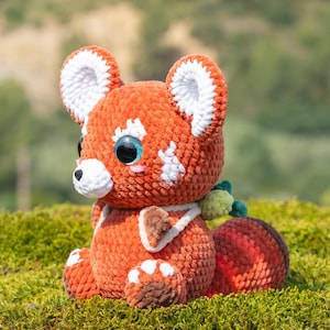 May include: A plush red panda toy made of soft orange and white yarn. The creature features large, glossy dark eyes, a white muzzle, and pointed ears with white inner details. It has a striped tail and a small green accessory attached to its back.