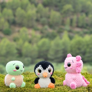 Pode incluir: Três pequenos peluches de crochê, incluindo uma tartaruga verde-clara com carapaça bege, um pinguim preto e branco com bico laranja e um axolote rosa. Cada brinquedo tem olhos pretos grandes e brilhantes e detalhes de bochechas rosas.