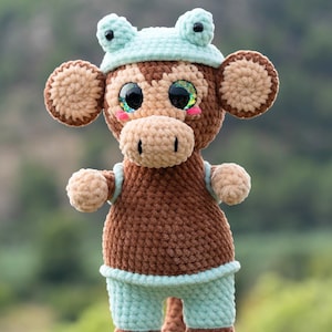 Puede incluir: Una muñeca de mono de ganchillo hecha a mano con un sombrero y pantalones cortos de rana. El peluche presenta hilo marrón y beige, ojos grandes y brillantes y detalles en azul claro. El mono tiene un cuerpo redondo, piernas y brazos largos.