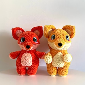 Könnte beinhalten: Zwei gehäkelte Fuchs-Plüschtiere, eines orange und eines gelb, mit blauen Augen und rosa Nasen.