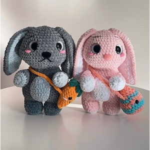 May include: Two crocheted bunny plushies, one gray and one pink, with embroidered eyes and noses. The gray bunny is holding a small orange carrot, and the pink bunny is holding a blue and white crocheted bag.