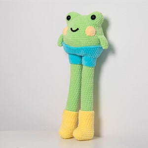 May include: A green crocheted frog toy with blue shorts and yellow boots. The frog has a friendly face with black eyes and a smile.
