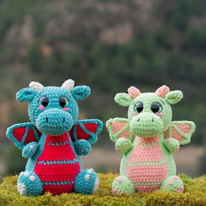 Puede incluir: Dos juguetes de peluche de dragón hechos a crochet. Uno es azul verdoso con alas rojas y vientre rosa. El otro es verde claro con alas rosas y vientre rosa. Ambos tienen ojos brillantes.
