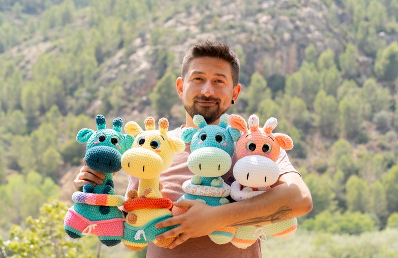 May include: A man is holding four crocheted giraffe toys. The giraffes are wearing colourful swimsuits and have large, bright eyes. The giraffes are different colours: teal, yellow, light blue, and white.