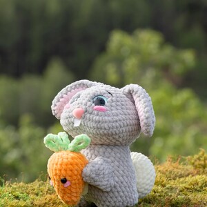 Puede incluir: Un juguete de conejo de crochet gris que sostiene una zanahoria de crochet naranja. El conejo tiene mejillas rosadas y ojos azules. La zanahoria tiene una parte superior de crochet verde.