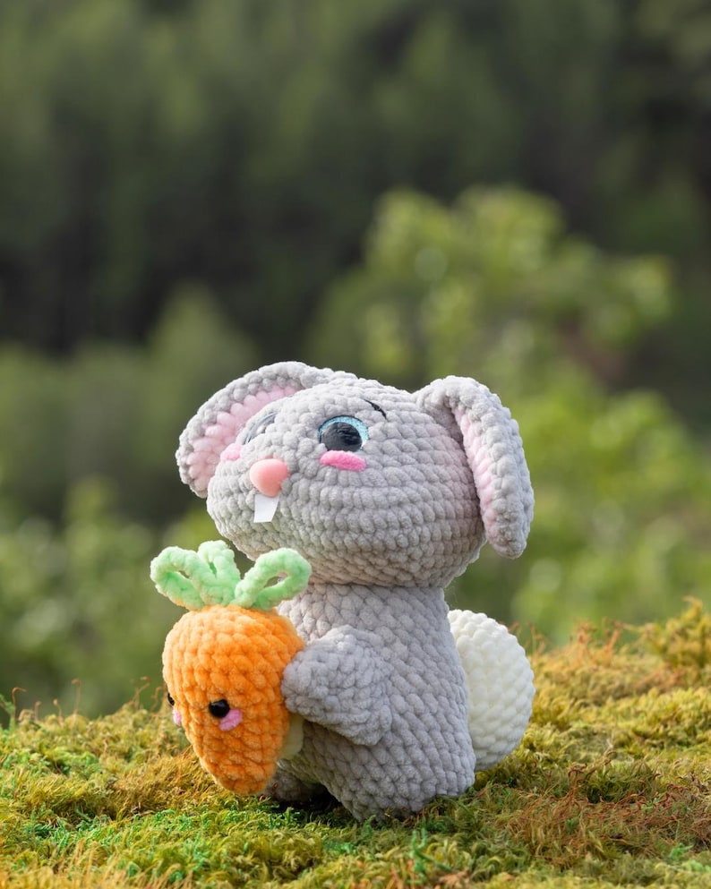 Puede incluir: Un juguete de conejo de crochet gris que sostiene una zanahoria de crochet naranja. El conejo tiene mejillas rosadas y ojos azules. La zanahoria tiene una parte superior de crochet verde.
