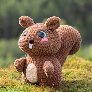 Puede incluir: Un juguete de peluche de ardilla de crochet marr&oacute;n con grandes ojos azules y una nariz rosa. El juguete est&aacute; sentado en un lecho de musgo verde.