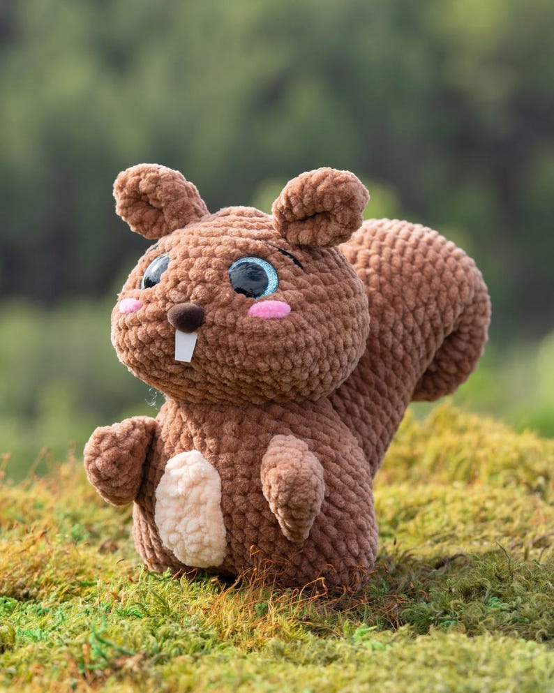 Puede incluir: Un juguete de peluche de ardilla de crochet marr&oacute;n con grandes ojos azules y una nariz rosa. El juguete est&aacute; sentado en un lecho de musgo verde.