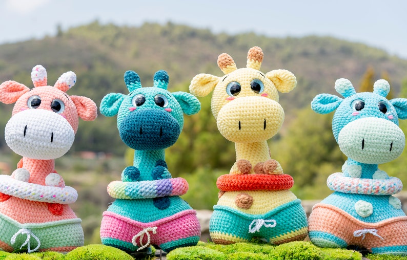May include: Four crocheted giraffe toys wearing colourful swimsuits and life preservers. The giraffes are in various colours, including pink, blue, yellow, and green.