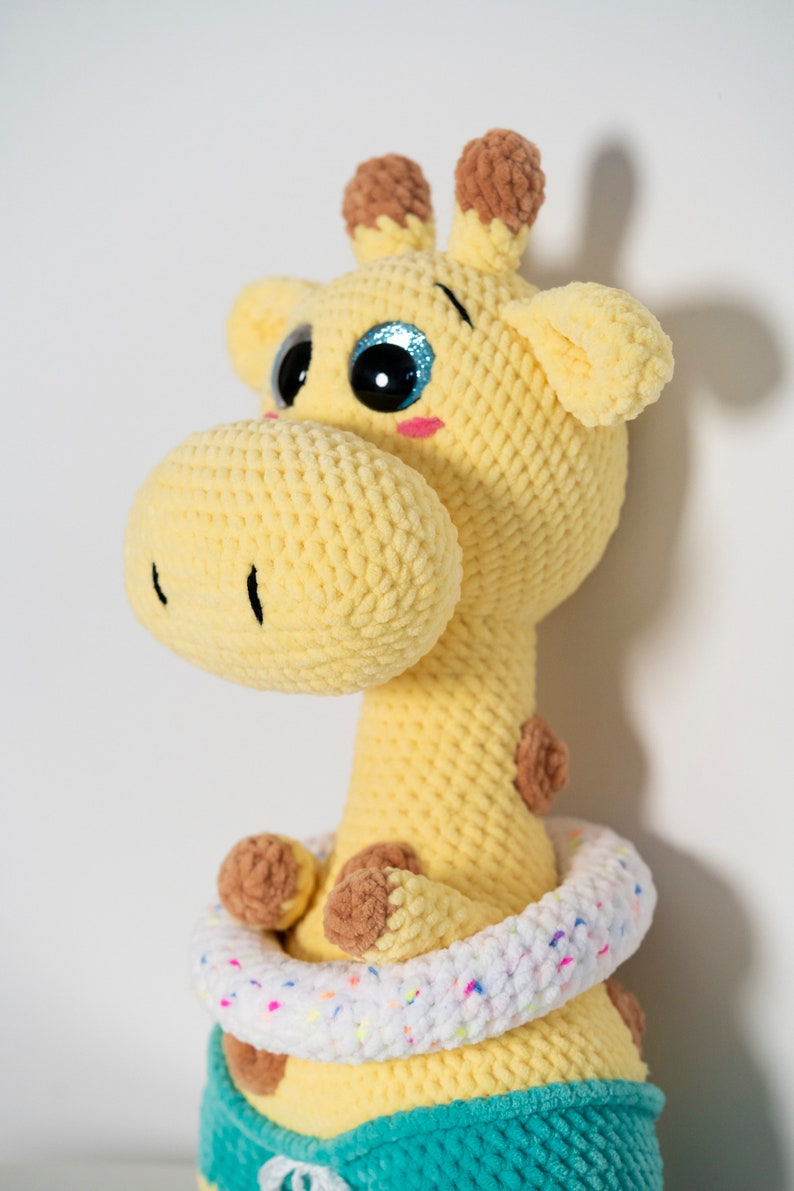 May include: A crocheted giraffe toy with large blue eyes and a white and pink ring around its neck. The giraffe is wearing a teal blue outfit.
