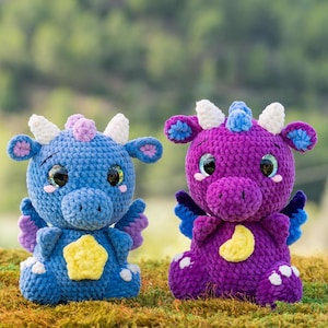 May include: Two handmade crochet dragon plush toys. One is blue with a yellow star on its belly, the other is purple with a yellow crescent moon. Both have large, sparkly eyes, white horns, and small wings.