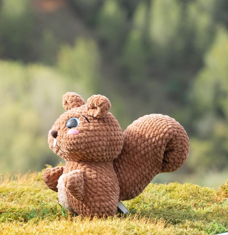 Puede incluir: Un juguete de peluche de ardilla de punto marr&oacute;n con ojos negros y mejillas rosadas. El juguete est&aacute; sentado en un lecho de musgo verde.