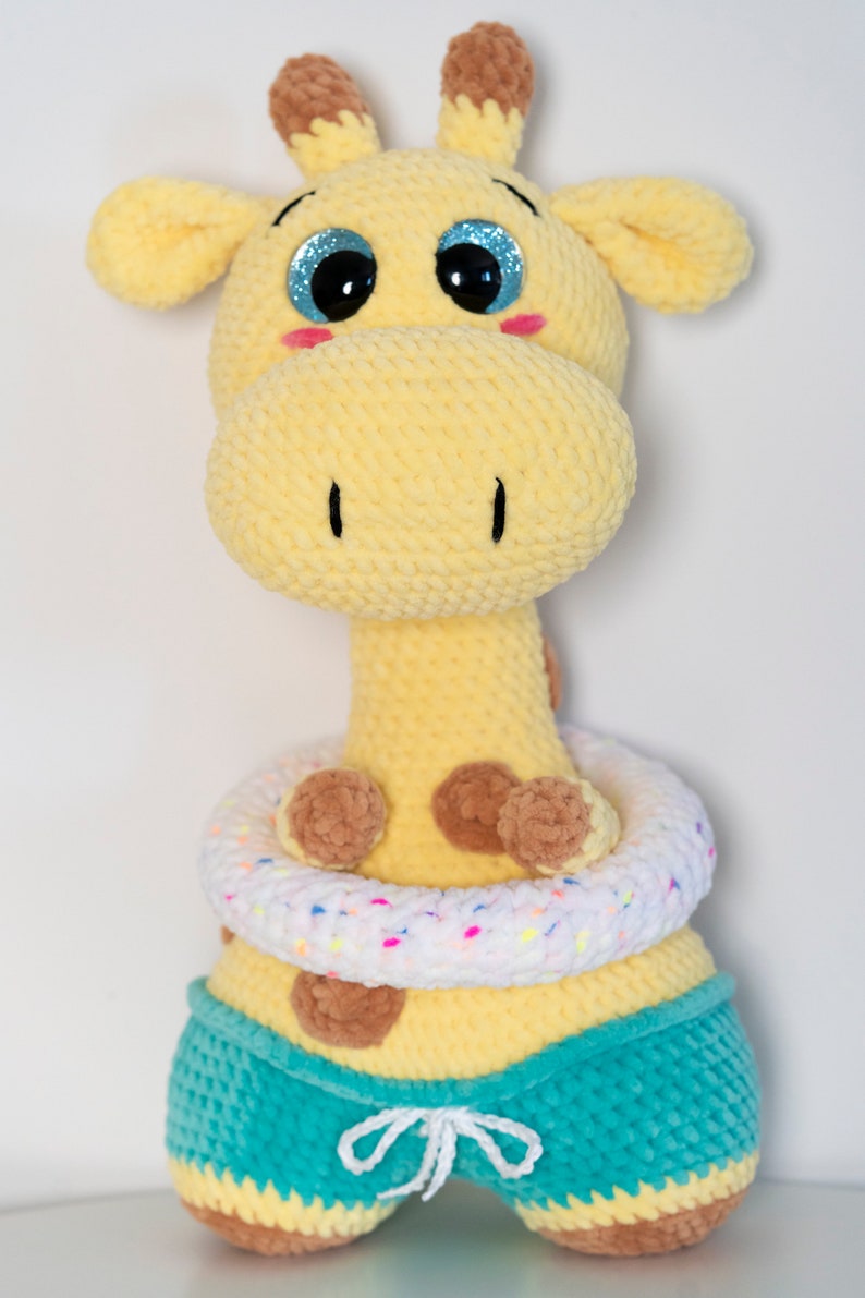 May include: A crocheted giraffe plush toy wearing a white and brown life preserver and teal blue swimming trunks. The giraffe has brown spots and blue eyes.