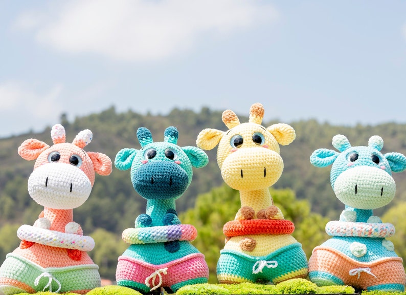 May include: Four crocheted giraffe toys wearing colourful swimsuits and life preservers. The giraffes are light blue, yellow, pink, and light blue with a rainbow pattern.