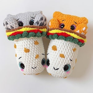May include: Two crocheted stuffed toys shaped like a hamburger and a hot dog. The hamburger is gray with a white bun and the hot dog is orange with a white bun. Both toys have smiling faces and blue eyes.