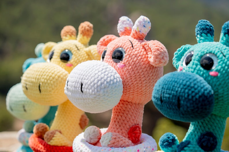 May include: Three crocheted giraffe plushies in yellow, pink and blue. Each plush has large, black eyes and a white nose. The yellow giraffe is wearing a red and white striped outfit. The pink giraffe is wearing a white and pink outfit. The blue giraffe is wearing a blue and white outfit.
