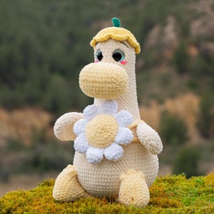 Puede incluir: Un juguete de peluche de crochet con forma de dinosaurio que lleva una flor amarilla y un sombrero amarillo con una hoja verde en la parte superior. El dinosaurio está sentado en un lecho de musgo verde.