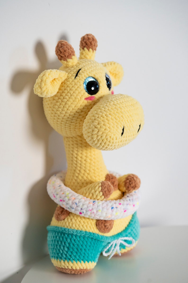 May include: A crocheted yellow giraffe wearing a white and pink inflatable ring and teal blue swimming trunks. The giraffe has brown ears and brown spots.