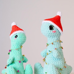 May include: Two crocheted dinosaur toys wearing Santa hats and decorated with colorful Christmas lights. The dinosaurs are light blue and have big, sparkly eyes.