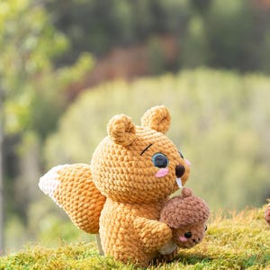 Puede incluir: Un juguete de peluche de ardilla de crochet con cola marr&oacute;n y blanca, sosteniendo un peque&ntilde;o juguete de peluche de bellota marr&oacute;n. La ardilla tiene la nariz rosa y los ojos negros.