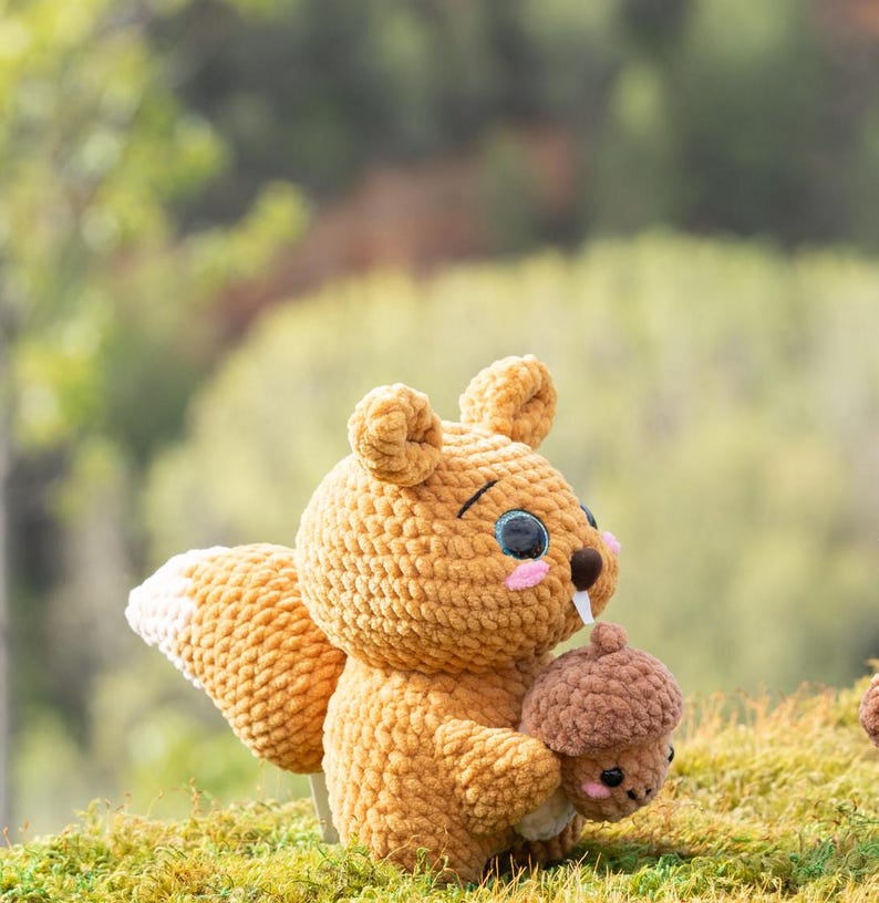 Puede incluir: Un juguete de peluche de ardilla de crochet con cola marr&oacute;n y blanca, sosteniendo un peque&ntilde;o juguete de peluche de bellota marr&oacute;n. La ardilla tiene la nariz rosa y los ojos negros.