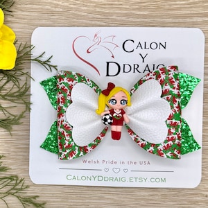 May include: A green and red glitter hair bow with a white heart-shaped center. A small figurine of a girl wearing a red and white soccer uniform and holding a soccer ball is attached to the center of the bow. The bow is on a white card with the text "Calon Y Ddraig" and "Welsh Pride in the USA" in black text. The text "CALONYDDRAIG.ETSY.COM" is also on the card.
