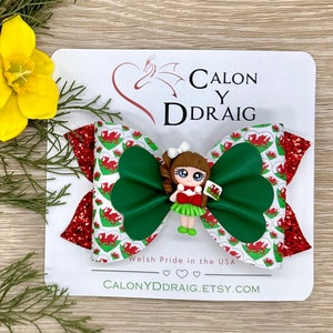 May include: A green and red bow with a white background featuring a red dragon with a white heart. The bow has a small doll with brown hair and a red dress holding a Welsh flag. The text on the card reads "Calon Y Ddraig" and "Welsh Pride in the USA".