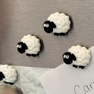 May include: Four white and black sheep magnets with black eyes. The magnets are attached to a gray surface.