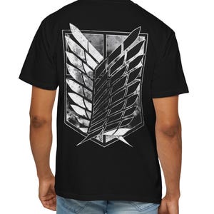Puede incluir: Una camiseta negra de manga corta con un gran gráfico blanco desgastado en la espalda. El diseño muestra el emblema de las Alas de la Libertad, que consiste en dos alas estilizadas y superpuestas dentro de un borde con forma de escudo texturizado.