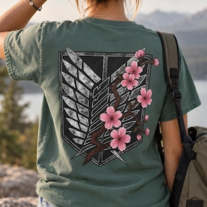 Può includere: Una t-shirt a maniche corte color verde salvia con una grande grafica sul retro. Il design mostra un emblema a scudo grigio scuro con motivi ad ali bianche, sovrapposto a un ramo marrone decorato con fiori di ciliegio rosa brillante.
