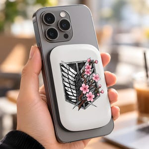 May include: A dark grey smartphone with a white magnetic battery pack attached to the back. The battery pack features a black and white shield emblem with a pair of wings, overlaid with a brown branch bearing several pink cherry blossoms.