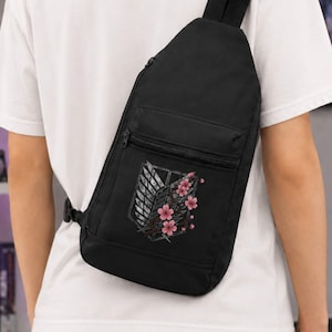 Puede incluir: Una mochila bandolera negra con un bolsillo frontal con cremallera que presenta un gráfico impreso de un emblema de escudo gris adornado con flores de cerezo rosas. La bolsa tiene una correa ajustable y se lleva sobre la espalda con una camiseta blanca.