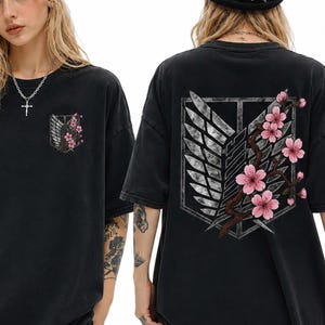 May include: Black short-sleeved t-shirt with a pocket featuring a floral design. The back of the shirt displays a large emblem with wings and a floral branch. The shirt has a vintage, worn look.