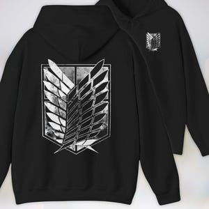 Puede incluir: Sudadera con capucha negra con un emblema blanco y gris en la parte delantera y una versión más pequeña en la capucha. El emblema representa alas estilizadas dentro de un escudo, con una estética vintage y desgastada. La sudadera tiene capucha con cordón y bolsillo canguro.