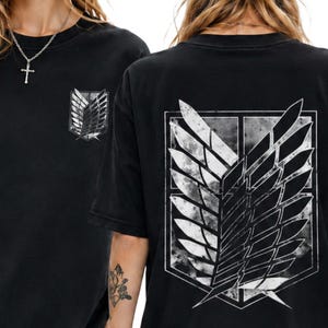 Op de afbeelding: Zwarte T-shirts met een wit en grijs embleem op de voor- en achterkant. Het embleem is een gestileerd vleugelontwerp in een schild. De shirts zijn gemaakt van een zacht materiaal en hebben een relaxte pasvorm.