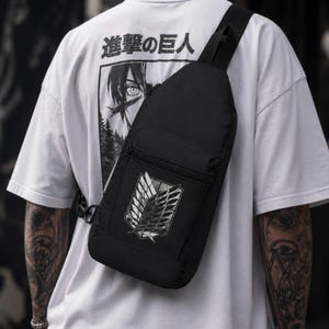 May include: A black sling backpack featuring a white graphic of the Wings of Freedom emblem from Attack on Titan on the front pocket. The bag has a single shoulder strap, a zippered main compartment, and a smaller zippered front pocket.