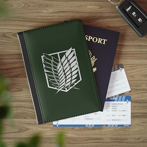 Può includere: Una custodia per passaporto verde scuro con l'emblema bianco delle Ali della Libertà, che copre parzialmente un passaporto blu scuro con scritte dorate. Due carte d'imbarco bianche e blu con dettagli del volo sono posizionate sotto gli oggetti.