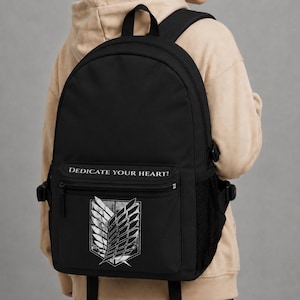 Può includere: Uno zaino nero con una grafica bianca dell'emblema delle Ali della Libertà sulla tasca anteriore. Sopra l'emblema, il testo DEDICATE YOUR HEART! è stampato in bianco. Lo zaino include una maniglia superiore, spallacci regolabili e tasche laterali in rete.