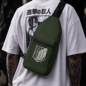 Peut inclure: Un sac à bandoulière vert olive arborant l'emblème blanc des Ailes de la Liberté imprimé sur la poche avant. Le sac est doté d'une fermeture éclair noire et d'une sangle réglable noire. Il est porté sur un t-shirt blanc avec du texte japonais.