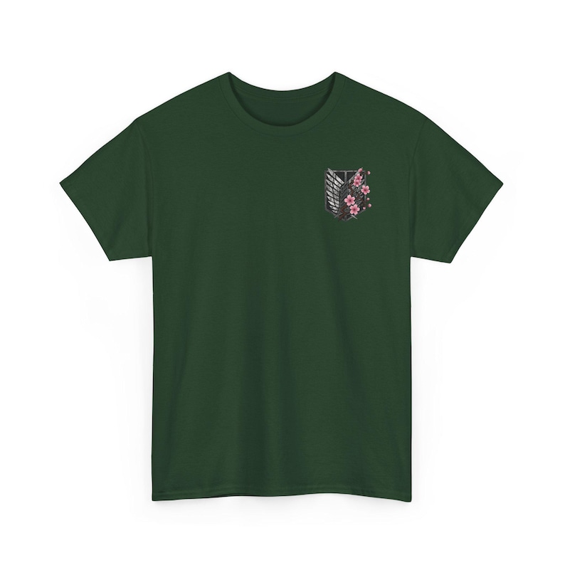 May include: Dark green t-shirt featuring a graphic of a winged emblem with cherry blossoms. The emblem is black and white with pink flowers. The shirt is a solid color and has short sleeves.