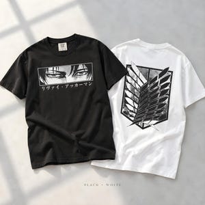 Könnte beinhalten: Ein schwarzes T-Shirt mit einer Grafik von Anime-Augen und dem japanischen Text "リヴァイ・アッカーマン" in einem rechteckigen Rahmen, kombiniert mit einem weißen T-Shirt, das auf der Rückseite ein großes schwarz-weißes "Wings of Freedom"-Emblem zeigt, beide mit kurzen Ärmeln und Rundhalsausschnitt.