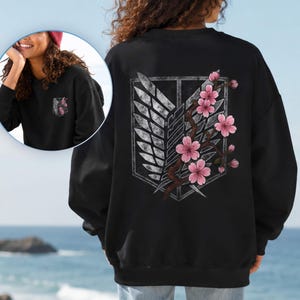 May include: A black long-sleeve sweatshirt featuring a large graphic on the back of a grey shield with wings and a branch of pink cherry blossoms. A smaller version of the same graphic is printed on the front left chest area of the garment.