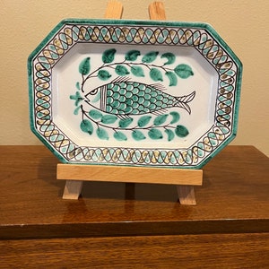 May include: A white ceramic serving dish with a green and gold border. The dish features a green fish with black details and green leaves on a white background.